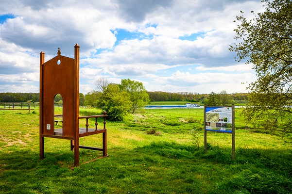 Een grote metalen stoel met uitzicht over het groene Maasdal, naast een informatiebord over het gebied.