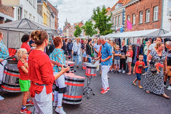Percussieoptreden op de Sjoemelmert in Roermond, met muzikanten in kleurrijke kleding en een enthousiast publiek tussen de marktkramen.
