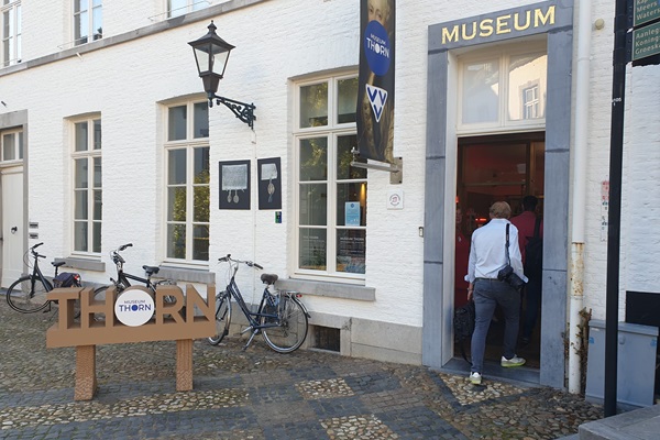 Entrance to Museum Thorn, located in a white historic building with an open door where visitors enter. In front of the building is a wooden sign reading ‘THORN’ and a logo. Some bicycles are parked against the wall, and a lantern hangs from the facade.