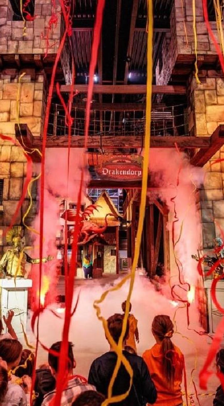 Kinderen en ouders vieren de opening van Drakendorp met slingers en rook bij een grote kasteelpoort.