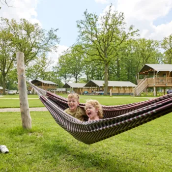 Twee kinderen lachen samen in een hangmat op een groen grasveld, met safaritenten op de achtergrond op een camping bij de Leistert.