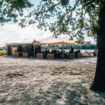 Terras van Het Blauwe Meertje aan het strand, met uitzicht over het water en beschutte zitplekken onder de bomen.